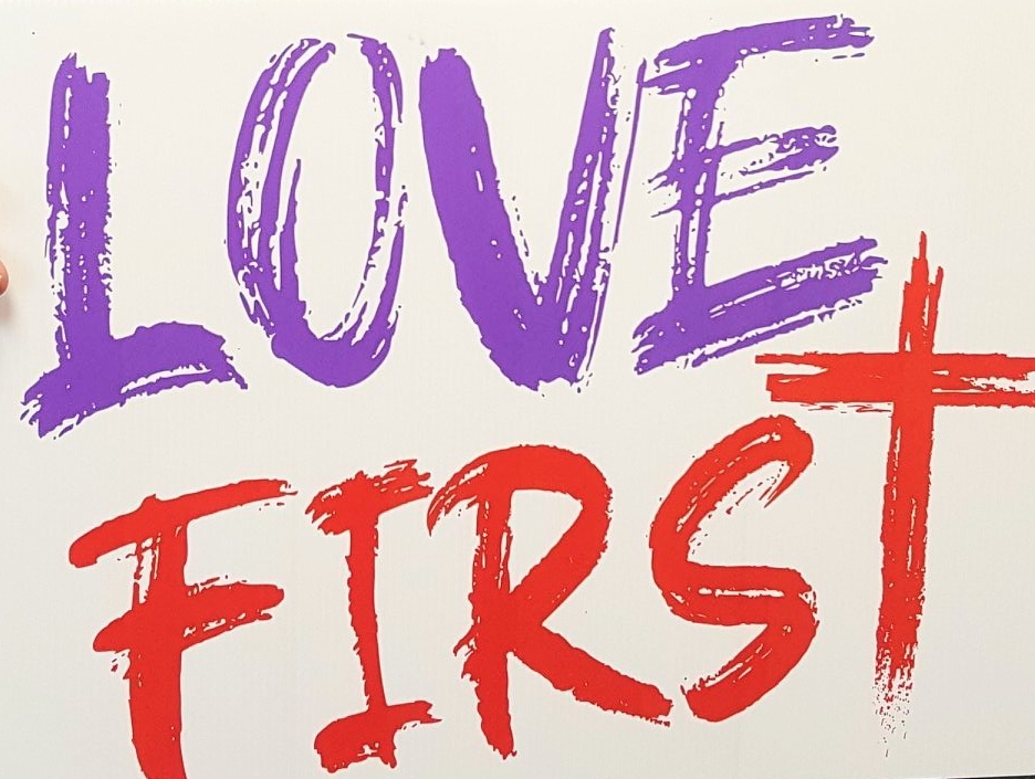 The words "Love First" in purple and red lettering with a paintbrush look